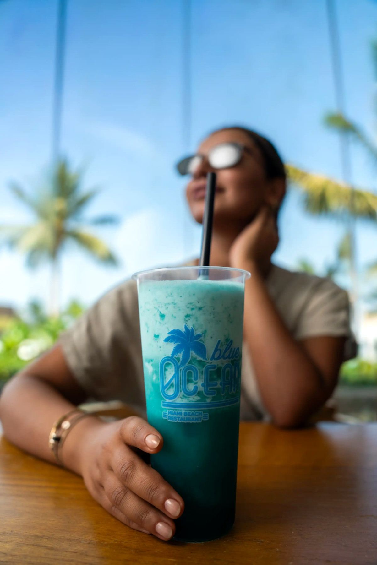 Iced blue mocktail held at a café table with palm trees behind at La Maison D’Obhur
