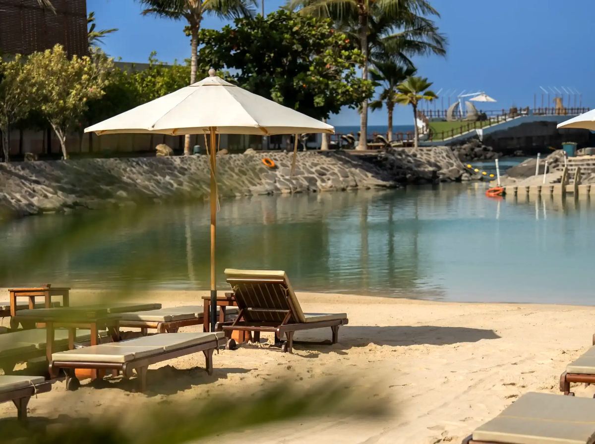 Wooden loungers and a cream parasol on the sand beside the calm lagoon at La Maison D’Obhur