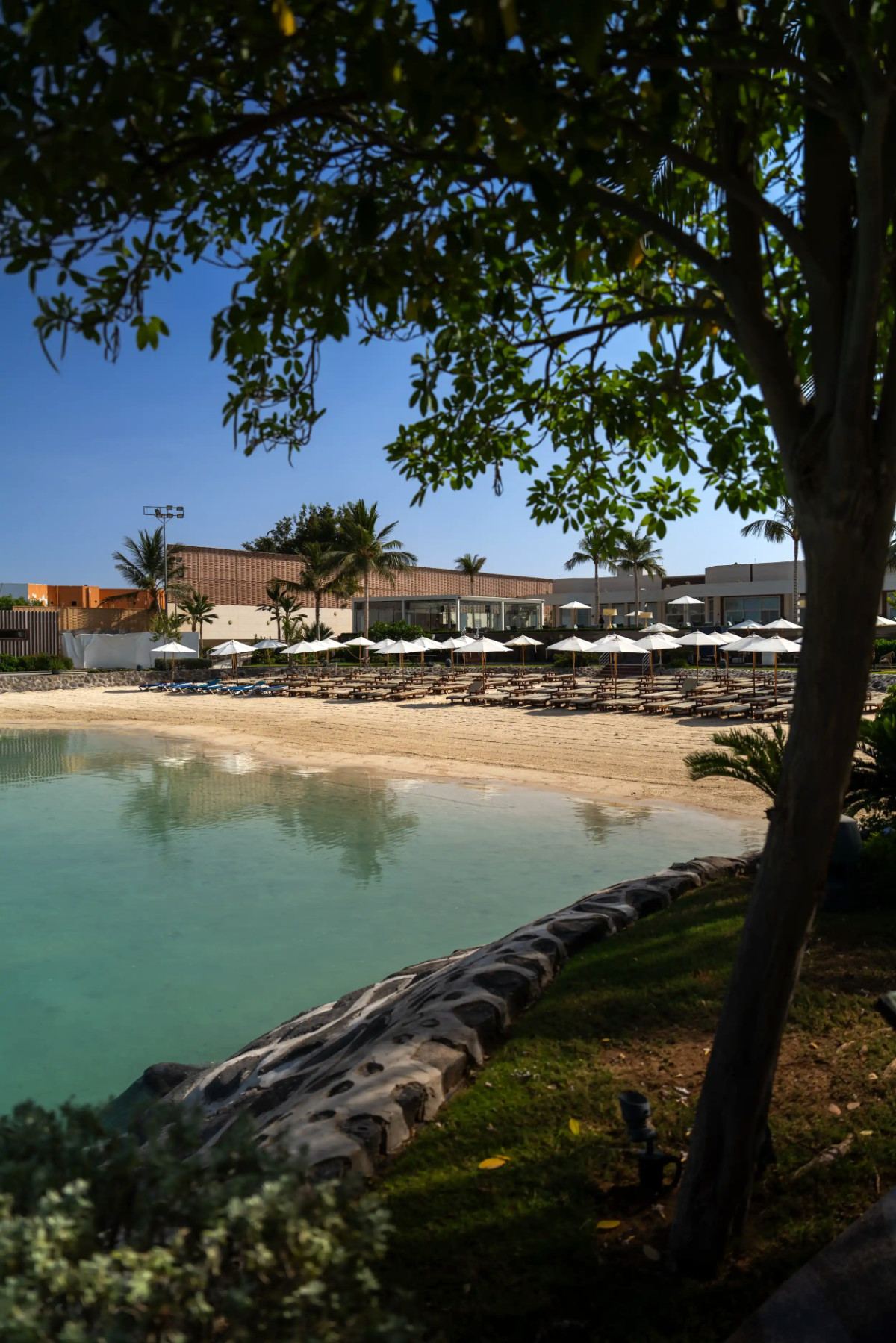Shady tree framing the calm lagoon and parasol-lined beach at La Maison D’Obhur