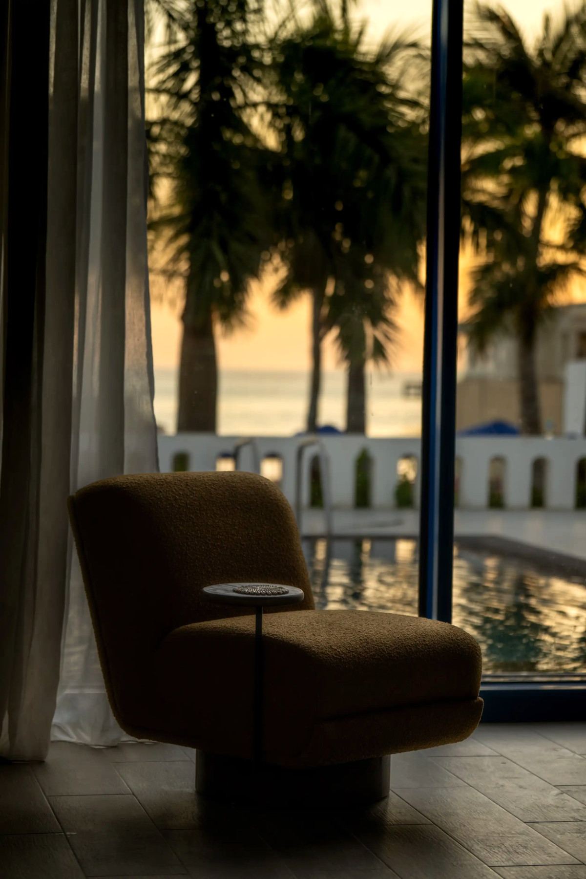 Lounge armchair by a window overlooking the pool, palms and sunset sea at Le Pont de l’Amitié
