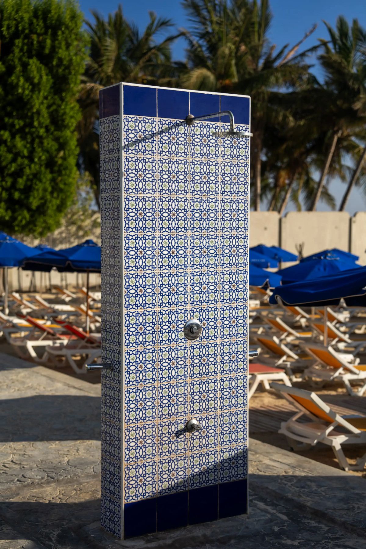 Blue geometric-tiled beach shower beside sunbeds and blue parasols at Le Pont de l’Amitié