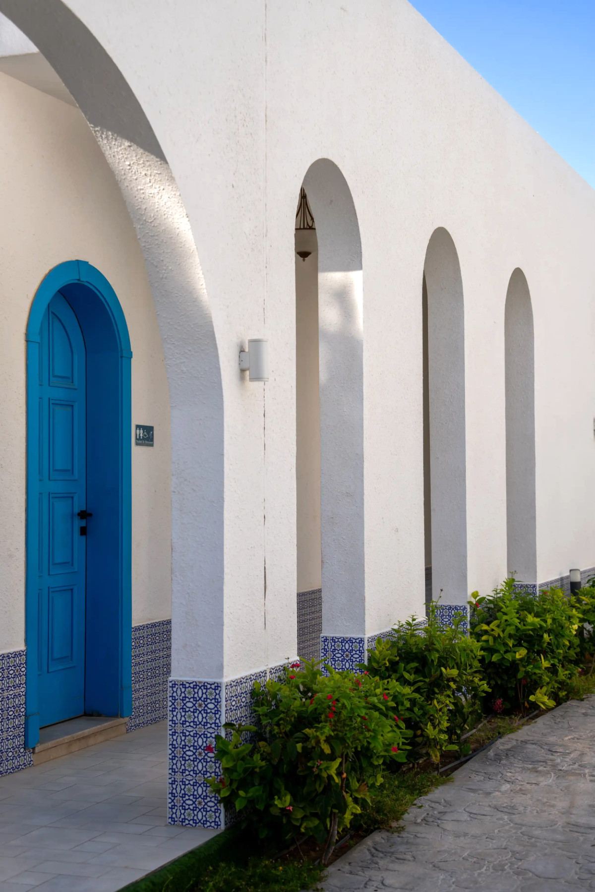 White arched colonnade with a blue door and patterned tile trim at Le Pont de l’Amitié