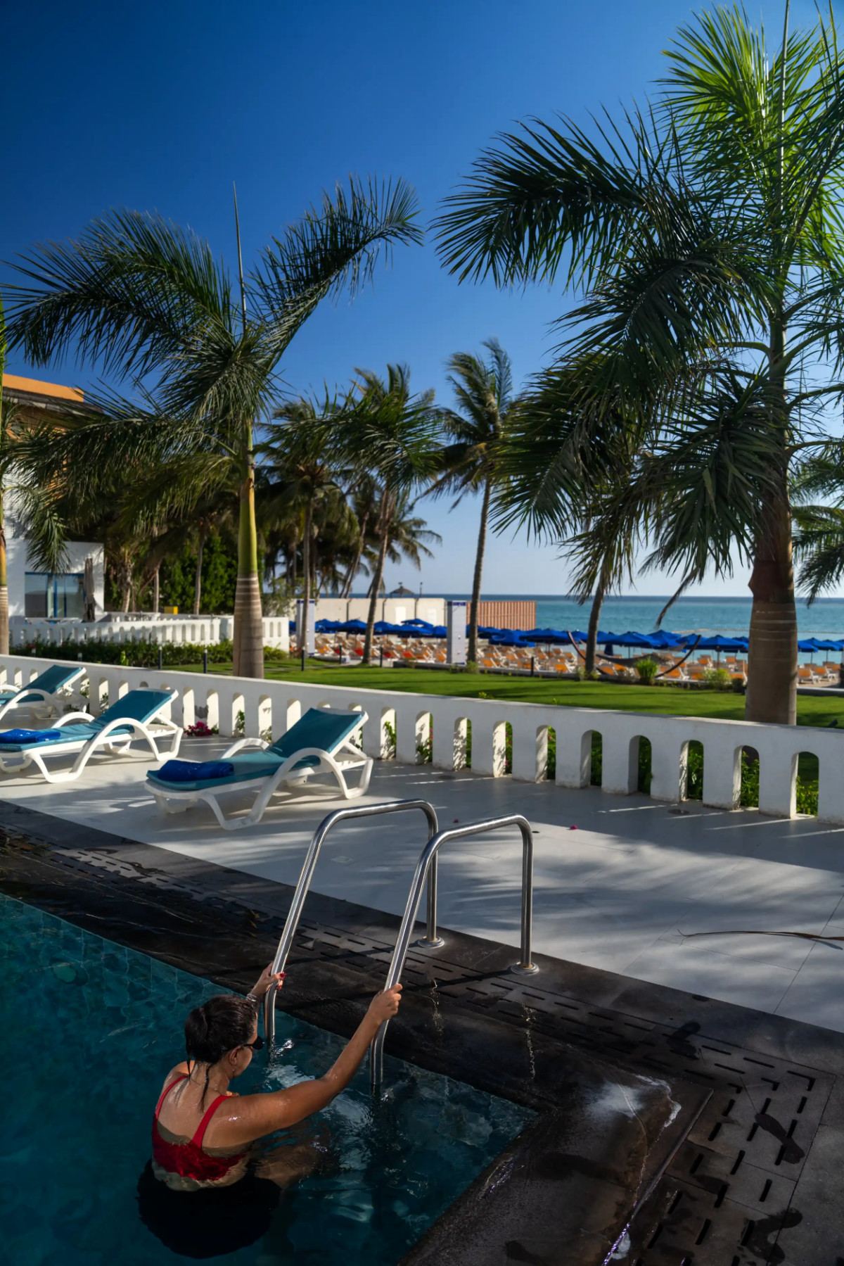 Swimmer at a pool ladder overlooking palms, sunbeds and the sea at Le Pont de l’Amitié