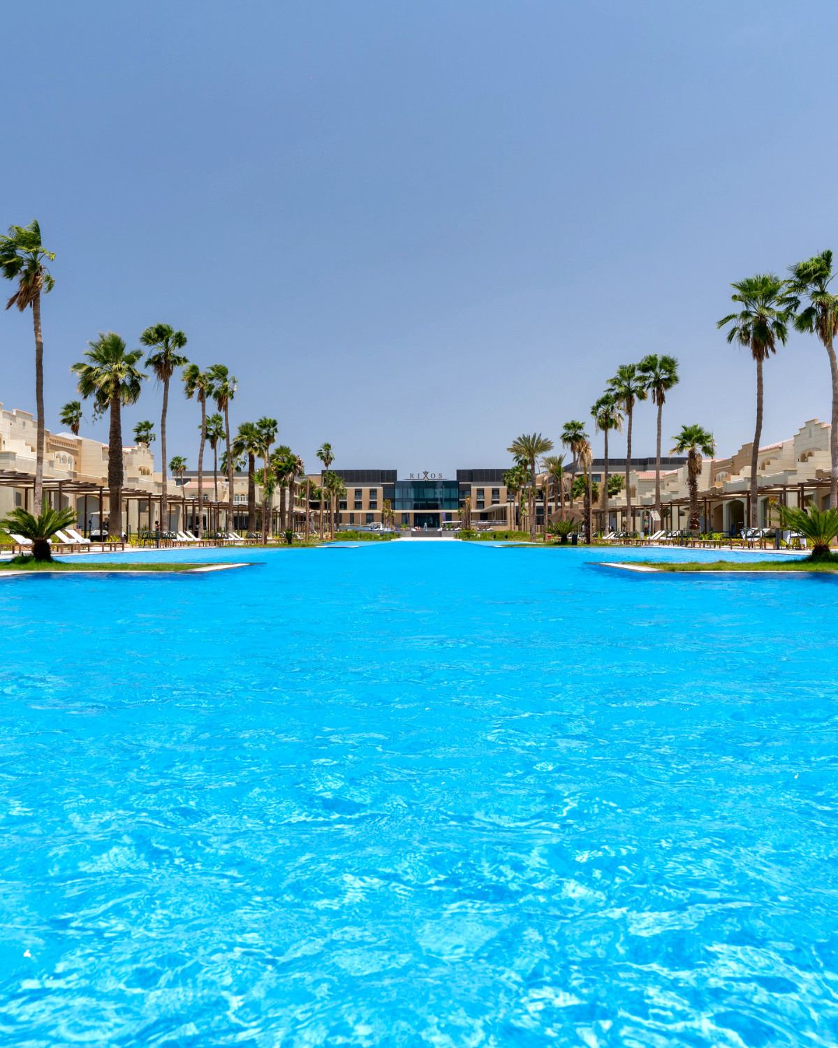 Turquoise pool at Rixos Obhur Jeddah framed by tall palms and the resort’s glass-fronted main building